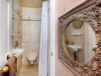 Cream-tiled bathroom with a white toilet, sink, curtains,& gold shower head & a gold framed mirror