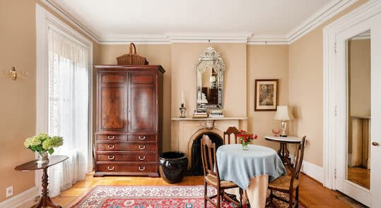 Bedroom with beige walls. Red area rug. Round Table with Chairs. Mirror above Fireplace Mantel. Tall Brown armoire.