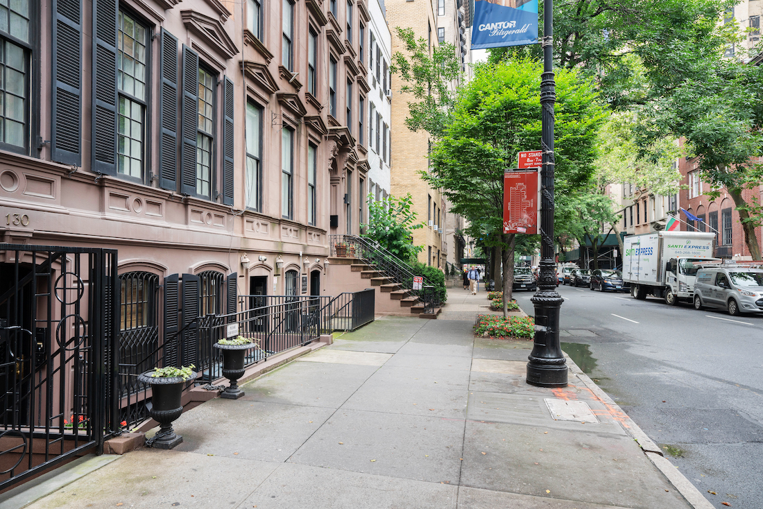 street scene with the brownstone building on the left. The small building is painted brown and has black shutters. Black lamp post and two black planters flanking the stairs. 