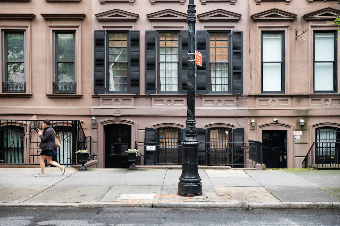 A brownstone building that is brown with black shutters. A lampost is in front and a girl walking. 