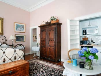 Interior room scene. Pink walls, iron-framed bed, tall wooden armoire, paintings on walls, large wooden chest at foot of bed, dark area rug.