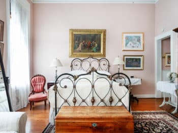 A well-lit bedroom with pastel pink walls, off-white bedding, dark decorative rug, antique iron bed frame, large print on bed wall with gold frame, and lace clad windows. Wooden chest in front of bed. Small pink antique chair.