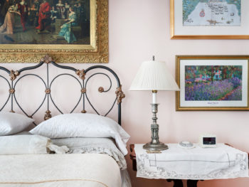 Room with pink walls, iron bed with scrolling, bedside table with white table cloth and silver lamp, prints on walls