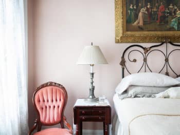 A well-lit bedroom with pastel pink walls, off-white bedding, dark decorative rug, Queen antique iron bed frame, large print on bed wall, and lace clad windows.