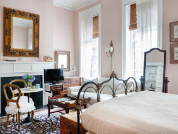 A well-lit bedroom with pastel pink walls, Fireplace , off-white bedding, dark decorative rug, antique iron bed frame, large print with gold frame on bed wall, and lace clad windows.