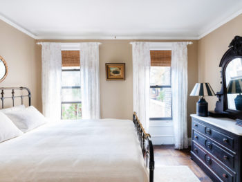 Large Back Bedroom Beige Walls, Iron and Brass Queen Bed, Rug, antique dresser, mirror behind bed. Lamp and painting