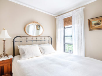 Back Bedroom with Beige Walls, Iron and Brass Queen Bed, Rug, antique dresser, mirror behind bed. Lamp and painting