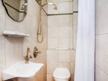 Cream-tiled bathroom with a white toilet, sink, curtains,& gold shower head & a mirror above sink