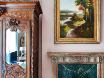 Bedroom with pink walls. Carved wood fireplace with green marble surround. Very large landscape painting with gold frame above fireplace.