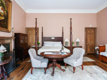 Spacious bedroom with four poster bed with white bedspread. Oval table and two upholstered chairs. Area rugs on floor.