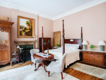 Bedroom with pale pink walls. Four poster bed and oval table at foot of bed. Area rugs on floor.