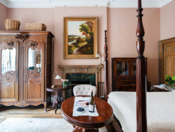 Spacious bedroom with pink walls. Four poster bed with white bedspread. Large painting on wall above fireplace.