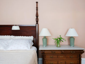 Corner of four poster bed with white bedspread and white pillows. Marble topped dresser with two green lamps.