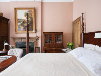 Spacious bedroom with pink walls. Four poster bed with white bedspread. Large Painting above fireplace.