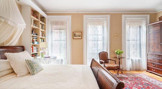 Bedroom with beige walls. Dark wood sleigh bed. White coverlet. 3 windows with white lace curtains. Red area rug.