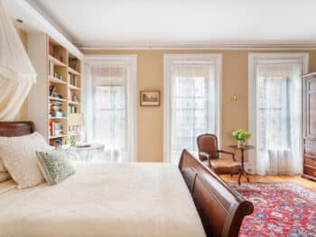 Room with beige walls, 3 windows with lace curtains and bed with white coverlet. Red Rugs.