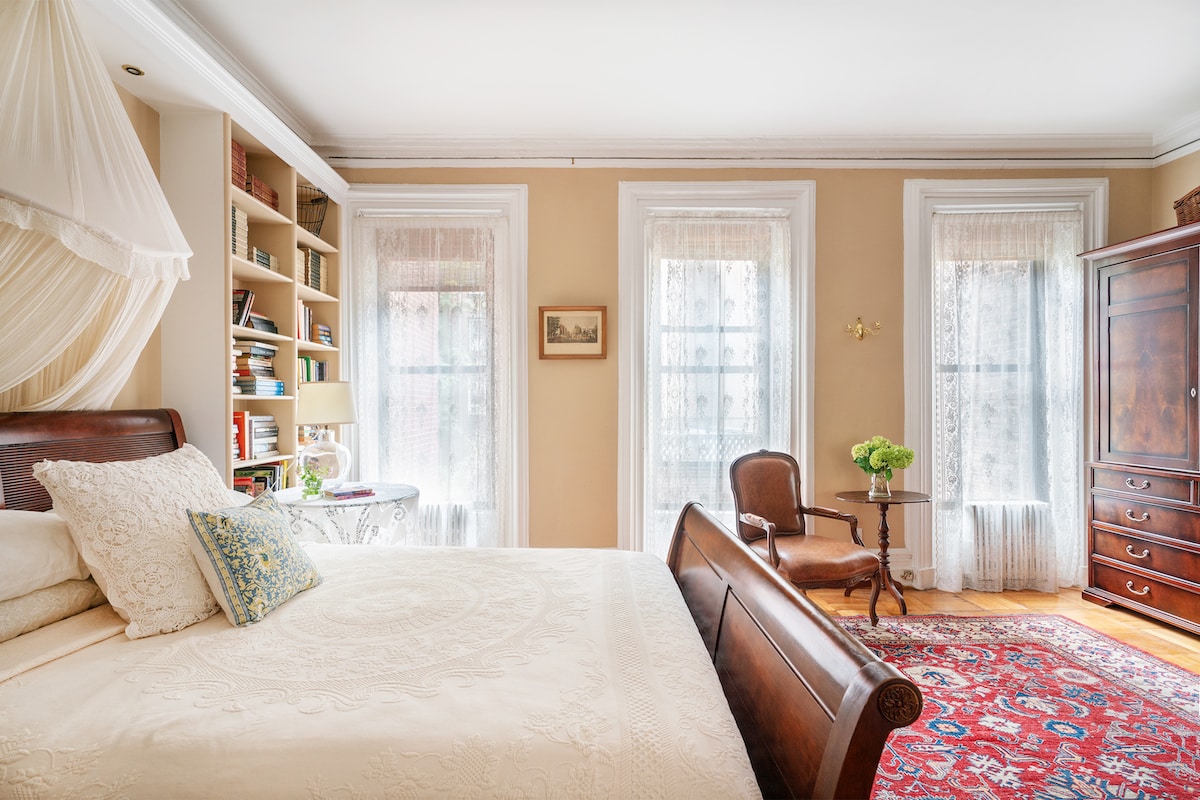 Room with beige walls, 3 windows with lace curtains and bed with white coverlet. Red Rugs.