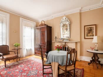 Room with Beige walls, brown wood armoire and table with blue table cloth and 3 chairs. Red Rugs and Mirror above fireplace