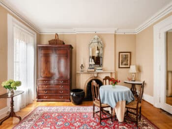 Room with Beige walls, brown wood armoire and table with blue table cloth and 3 chairs. Red Rugs and Mirror above fireplace