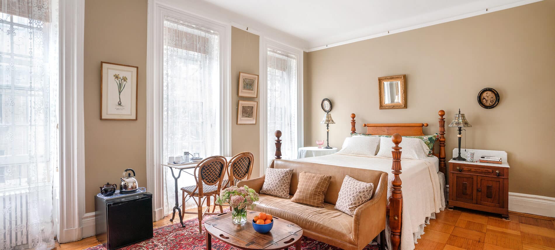 Bedroom with red rug. Bed with sofa at foot. White coverlet. Beige walls. 3 windows.