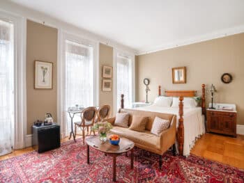 Bedroom with red rug. Bed with sofa at foot. White coverlet. Beige walls. 3 windows.