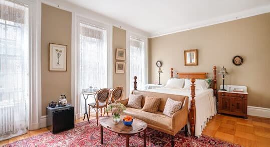 Bedroom with four poster wood bed, ivory bedding, leather love seat in front of bed and red area rug and 3 windows