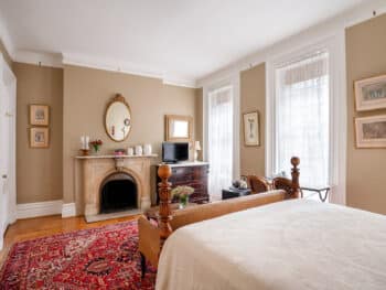 four poster bed with white cover. Red rug. Beige walls. Fireplace with mirror above.