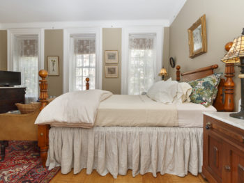 Side view of a beige-decorated bedroom with a wooden poster bed, three tall windows, & two bedstands