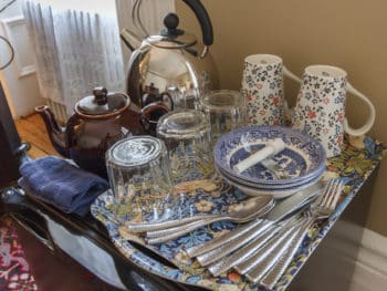 Tray with brown ceramic teapot, chrome electric kettle, teacups decorated with flowers, clear drinking glasses, silverware all on top of small fridge
