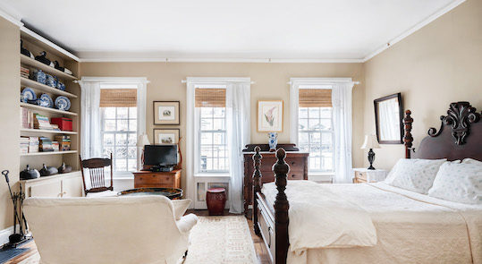 Bright bedroom furnished with a 4 poster bed, white love seat, wooden tv stand, fireplace, bookshelves and light colored area rug. 3 windows across the room.