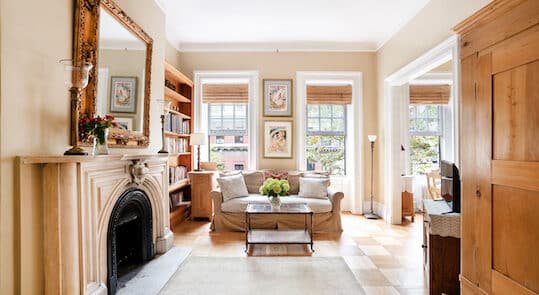 Brightly lit sitting area with a white & beige sofa, a glass coffee table, a marble fireplace with gilded mirror above, & two tall windows and light colored area rug over wood floors.