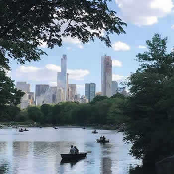 People row in boats on the Central Park Pond with trees & New York City's cityscape in the background