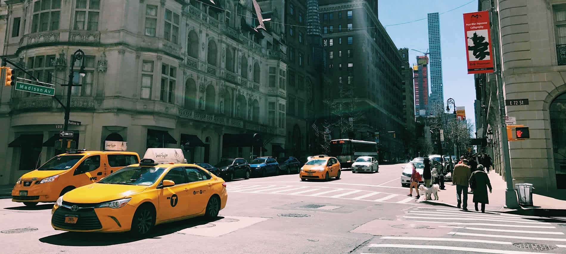 An intersection in New York City with multiple yellow taxis and crossing pedestrians during the day