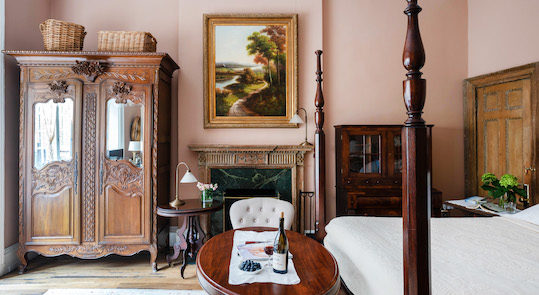 A well-lit room with pale rose-colored walls, a 4 poster bed, oval wooden table & chairs, tall wardrobe with mirrored doors, & a fireplace with large painting above.